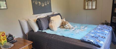 Een slaapkamer met een bed, kussens, een deken, een houten nachtkastje, een wandklok en een wandtapijt.