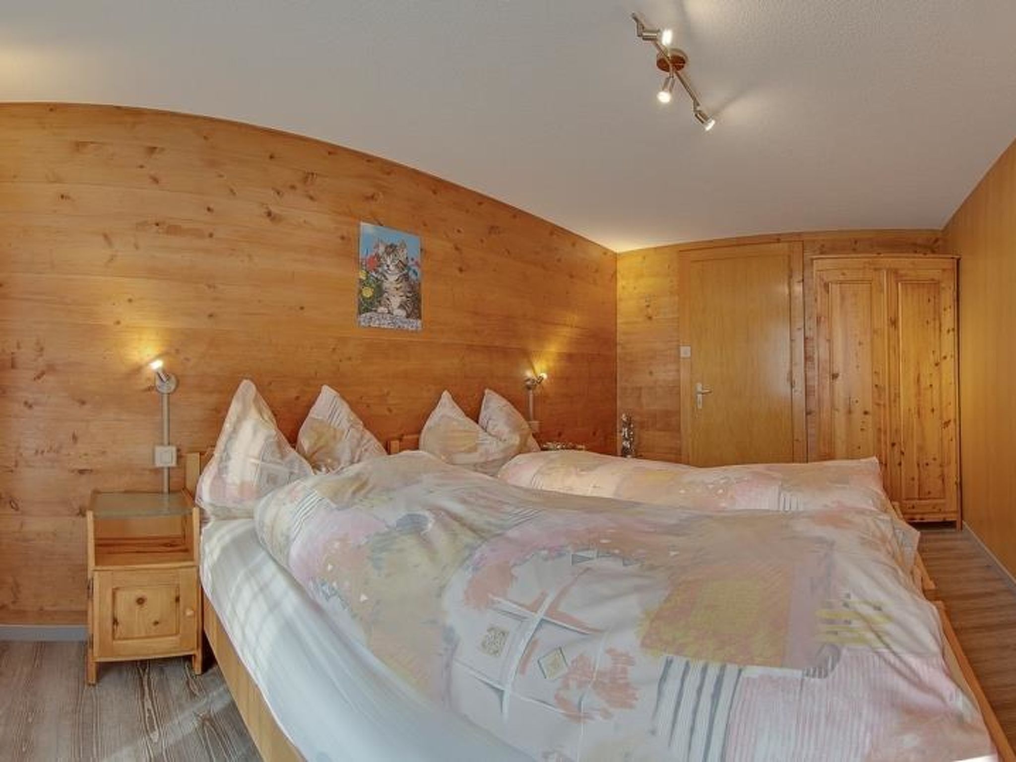 Ein Schlafzimmer mit Holzwand, einem Bett mit floralem Überwurf, zwei Nachttischlampen und einem Holzschrank.