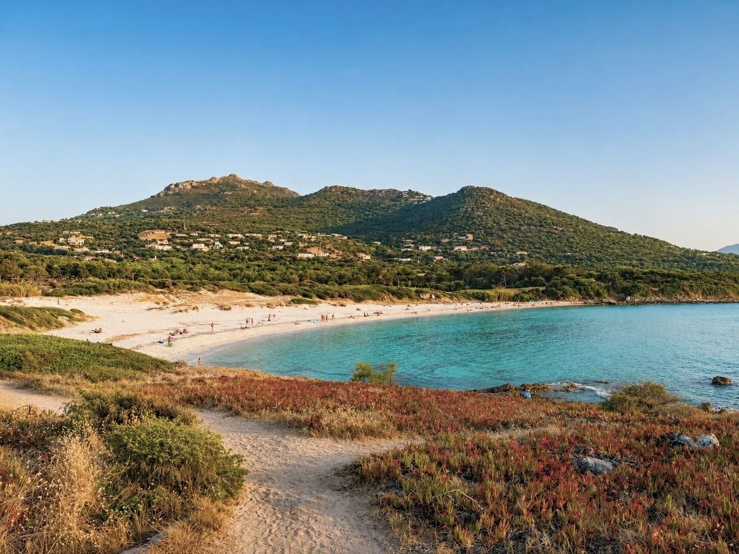 A coastal landscape with a sandy beach, clear turquoise waters, and a hilly backdrop with scattered buildings.