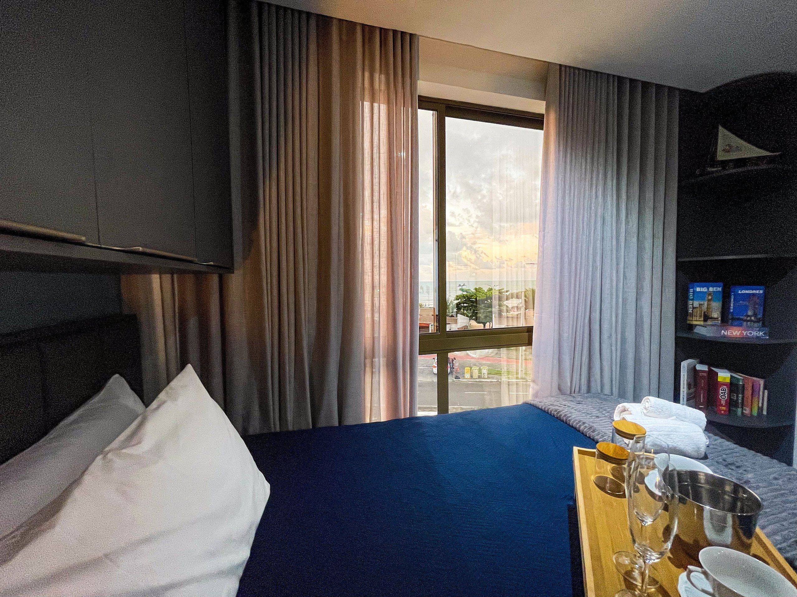 Quarto de hotel com cama, televisão, janela com cortinas, estante de livros e bandeja com copos e garrafa.