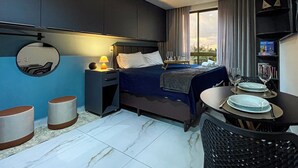 Habitación de hotel con cama, mesita de noche con lámpara, espejo redondo, pequeña mesa con platos y vasos, y vista al exterior a través de la ventana.