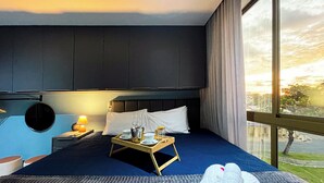 Habitación de hotel con una cama grande, una mesita de noche con lámpara, una bandeja con vasos y jarrón, y vistas al exterior.