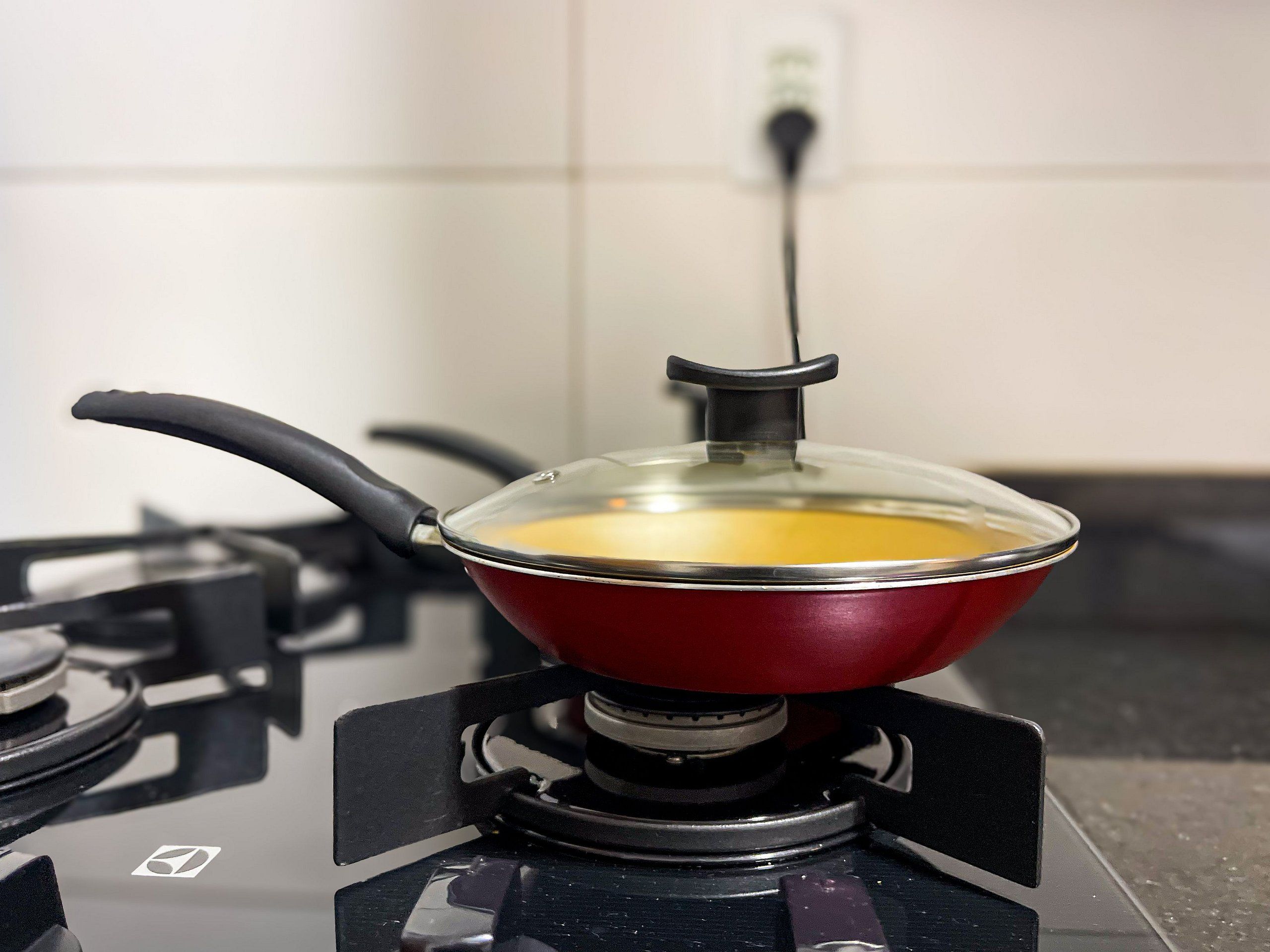 Een rode pan op een gasfornuis met een glazen deksel, een stopcontact en een zwart kookoppervlak.