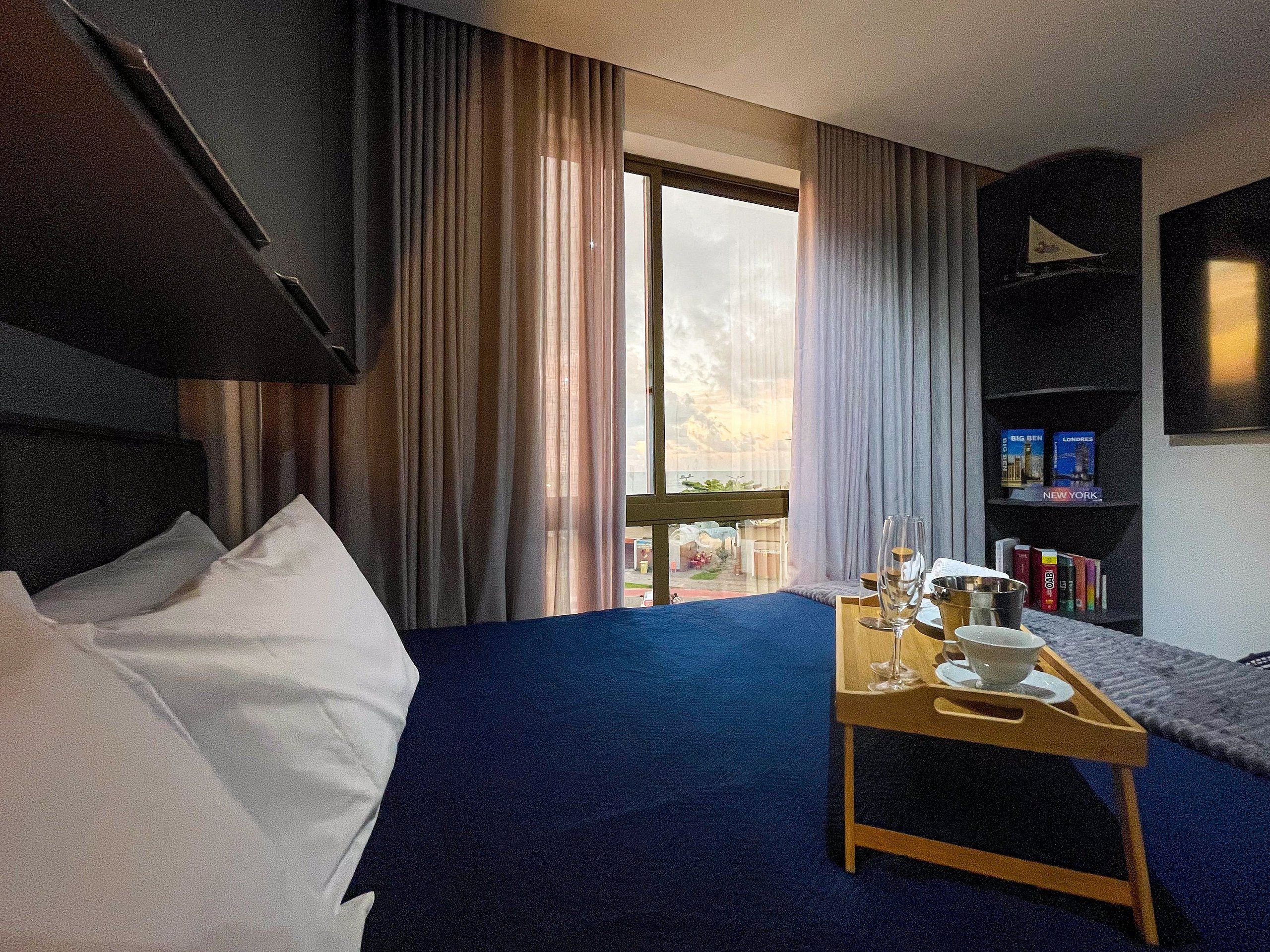 Quarto de hotel com cama, mesa de cabeceira de madeira com bandeja, estante de livros e vista para o exterior.