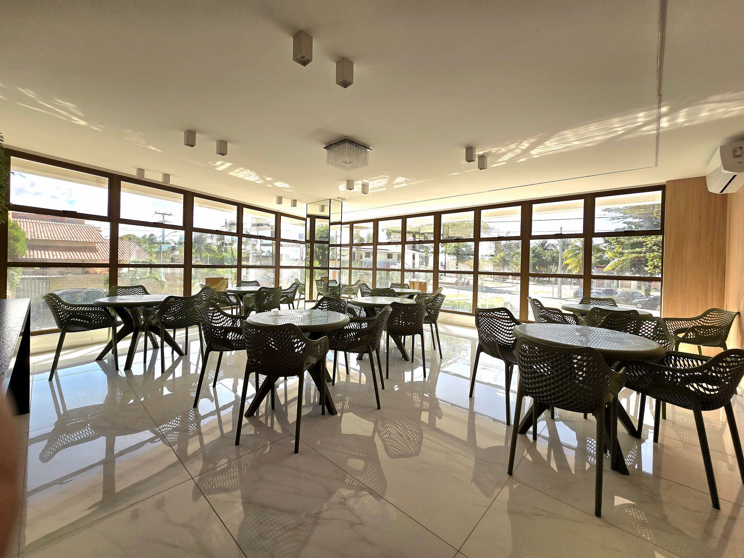A spacious indoor area with multiple tables and chairs, large windows offering a view of the outside, and a ceiling with recessed lighting.