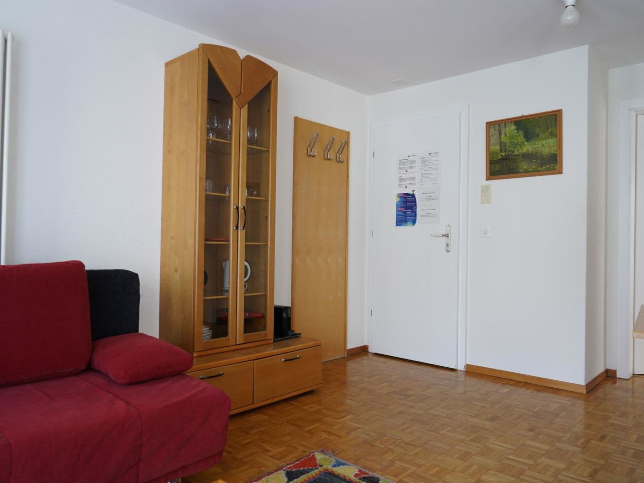A room with a red sofa, a wooden wardrobe with glass doors, a white door with a picture, and a wooden floor.