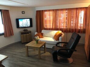 Ein Wohnzimmer mit einer Couch, einem schwarzen Sessel, einem Holztisch, einem Fernseher an der Wand und einem Fenster mit Vorhängen.