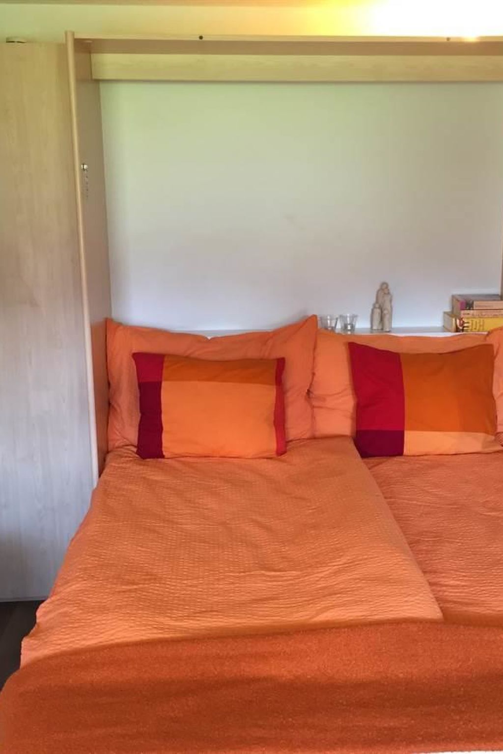 Un lit avec une couvre-lit et des oreillers orange, une petite étagère avec des livres et une figurine, et un mur sobre.