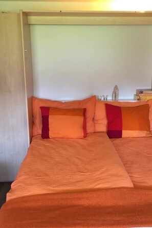 Een bed met een oranje sprei en kussens, een klein plankje met boeken en een beeldje, en een effen muur.