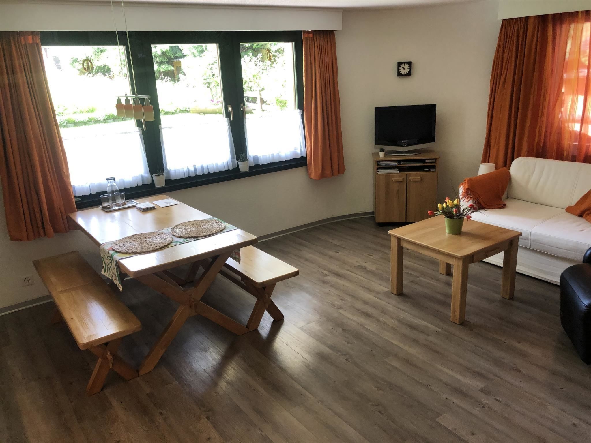A living room with a wooden floor, a sofa, a TV stand, and a dining table with benches.