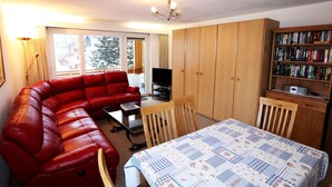 Una habitación amoblada con un sofá rojo, una mesa de comedor con mantel acolchado, un televisor, armarios de madera y una estantería.