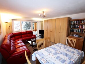 Ein möbliertes Zimmer mit einem roten Sofa, einem Esstisch mit einer gesteppten Tischdecke, einem Fernseher, Holzschränken und einem Bücherregal.