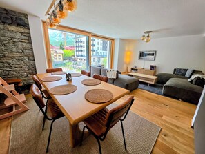 Ein modernes Wohnzimmer mit Essbereich, Steinwand, großen Fenstern und Blick auf Gebäude.