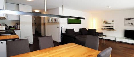 A modern kitchen with white cabinets, a wooden dining table, and a black sofa in the living area.