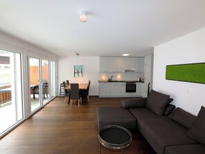 Ein modernes Wohnzimmer mit Sofa, Esstisch mit Stühlen, einer Küche mit Schränken und einer Schiebetür zum Außenbereich.