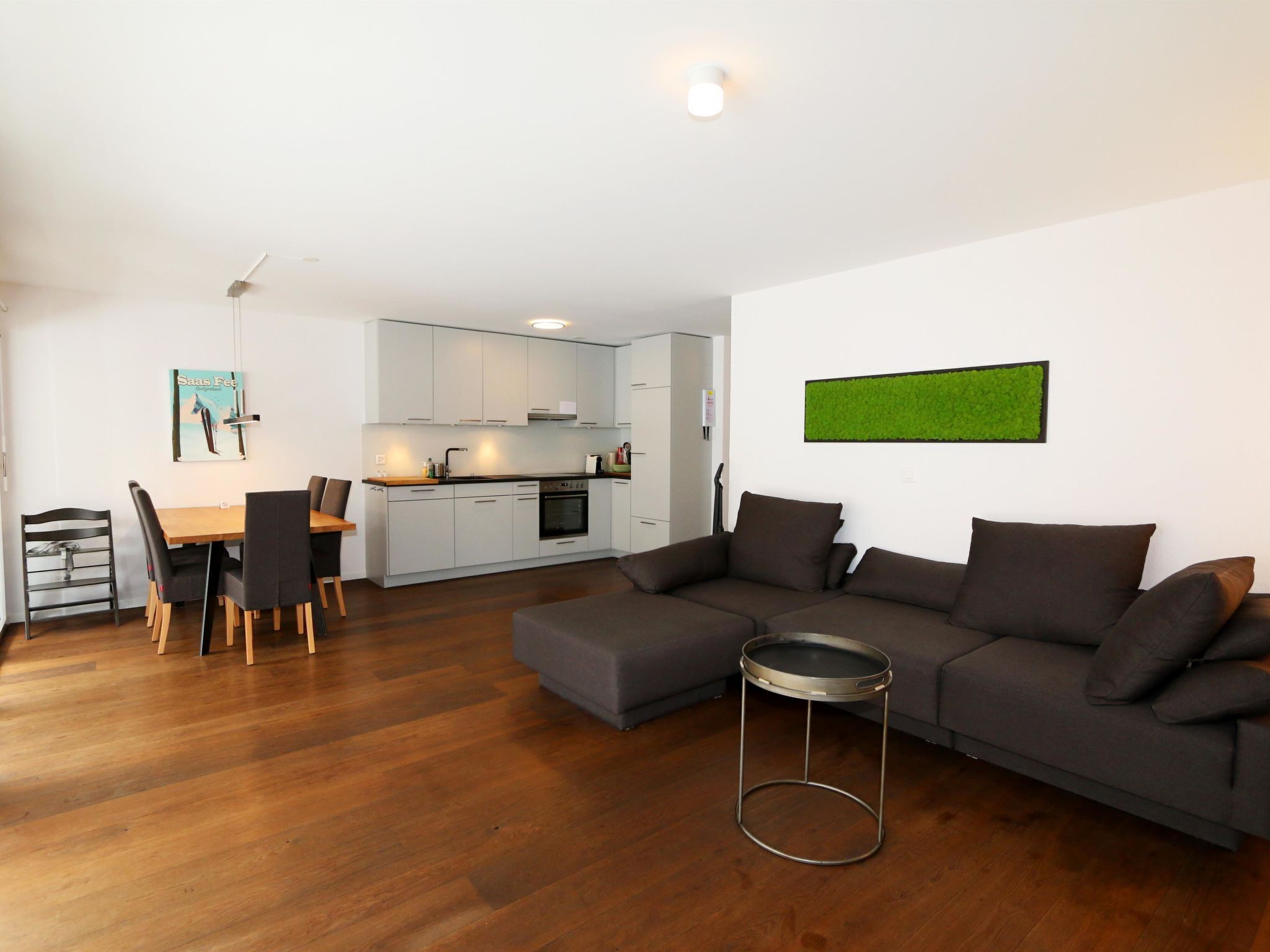 A modern living room with a grey sofa, a dining table with chairs, and a kitchen in the background.