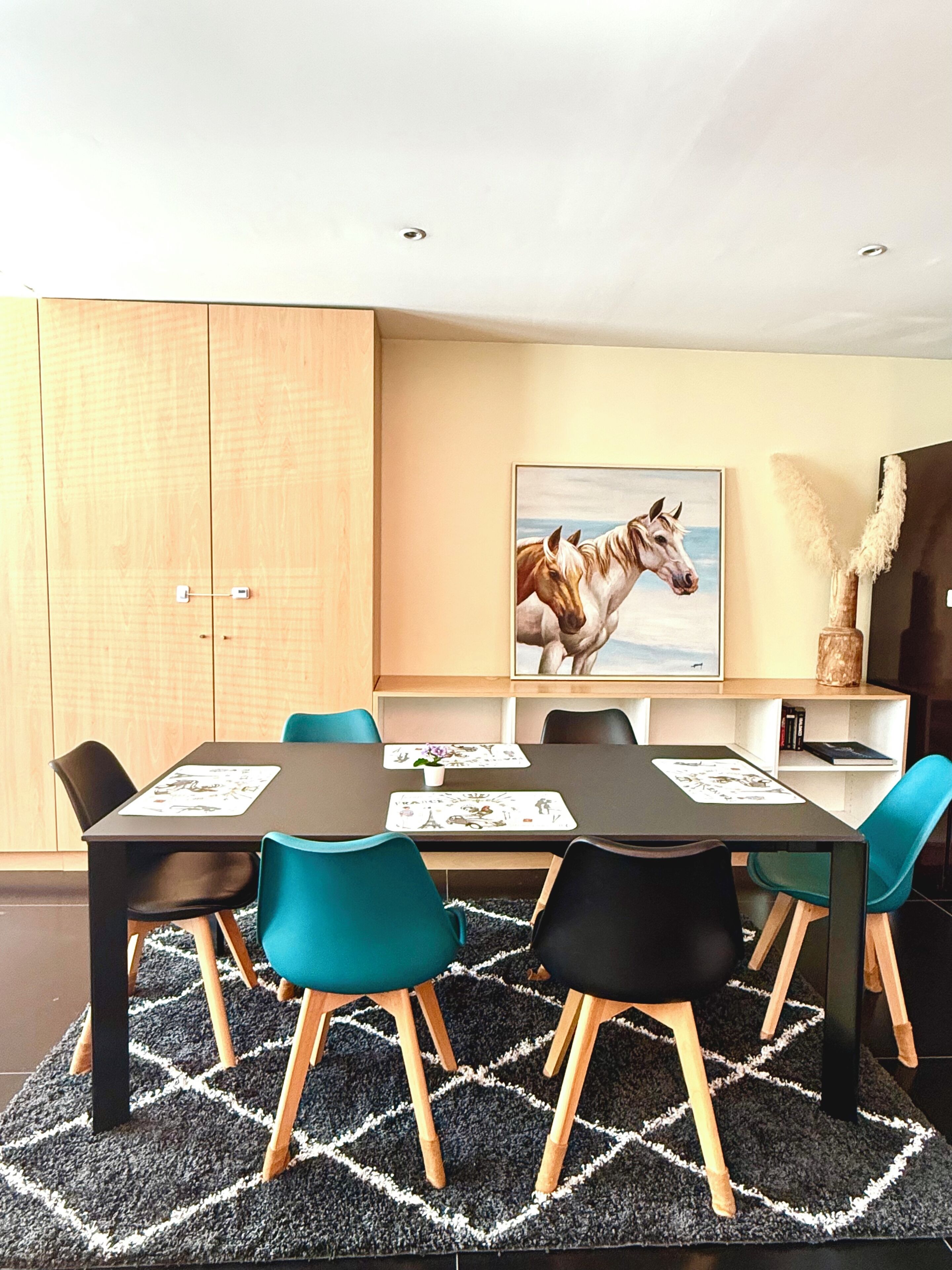 Un coin repas avec une table dressée pour quatre personnes, des chaises, un tapis et un tableau représentant des chevaux accroché au mur.
