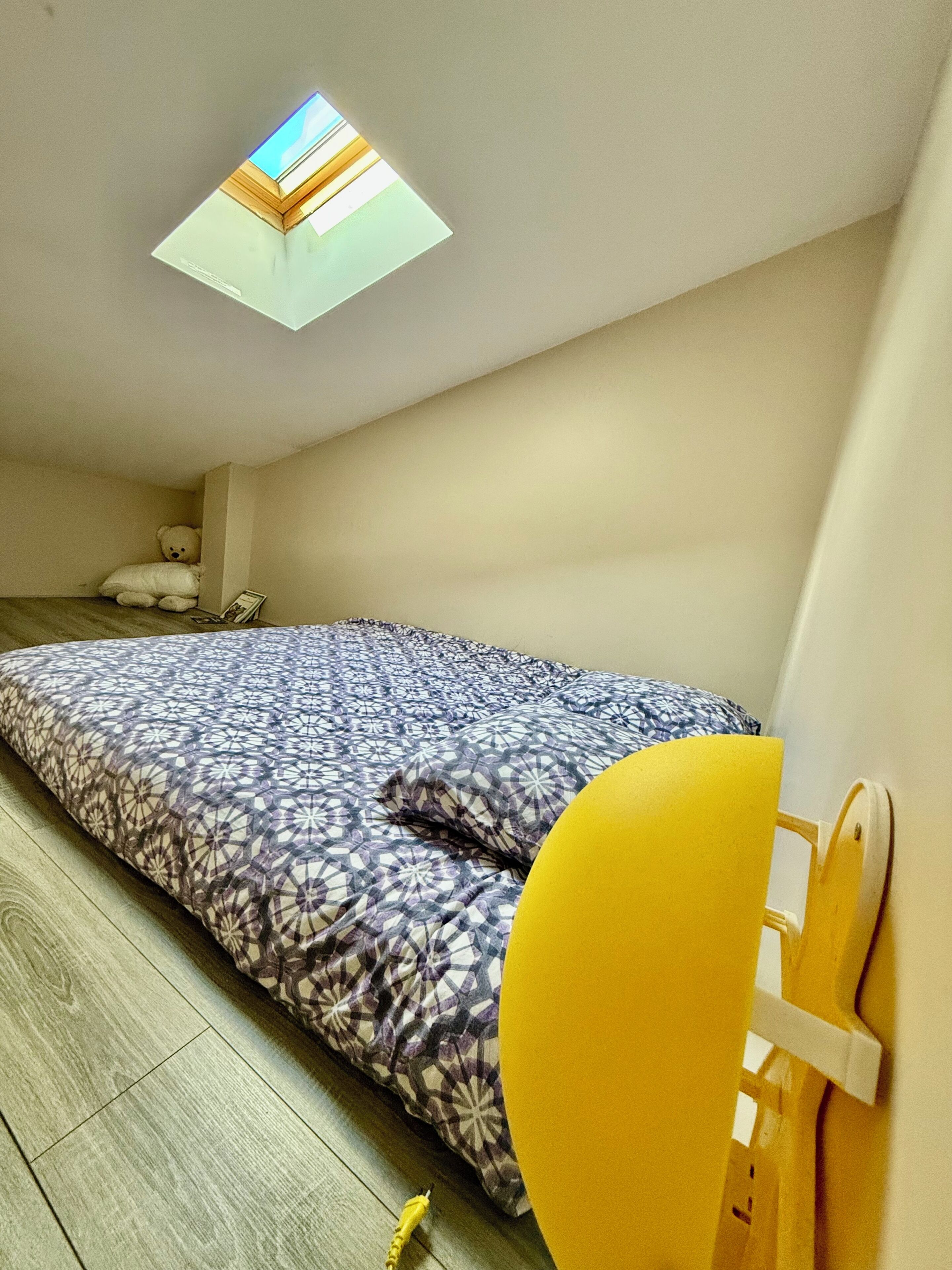 Ein Schlafzimmer mit einem Bett, einem gelben Sessel, einem Teddybären und einem Dachfenster.