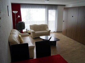 Uma sala de estar com dois sofás, uma mesa de centro, uma televisão e uma janela grande com cortinas.