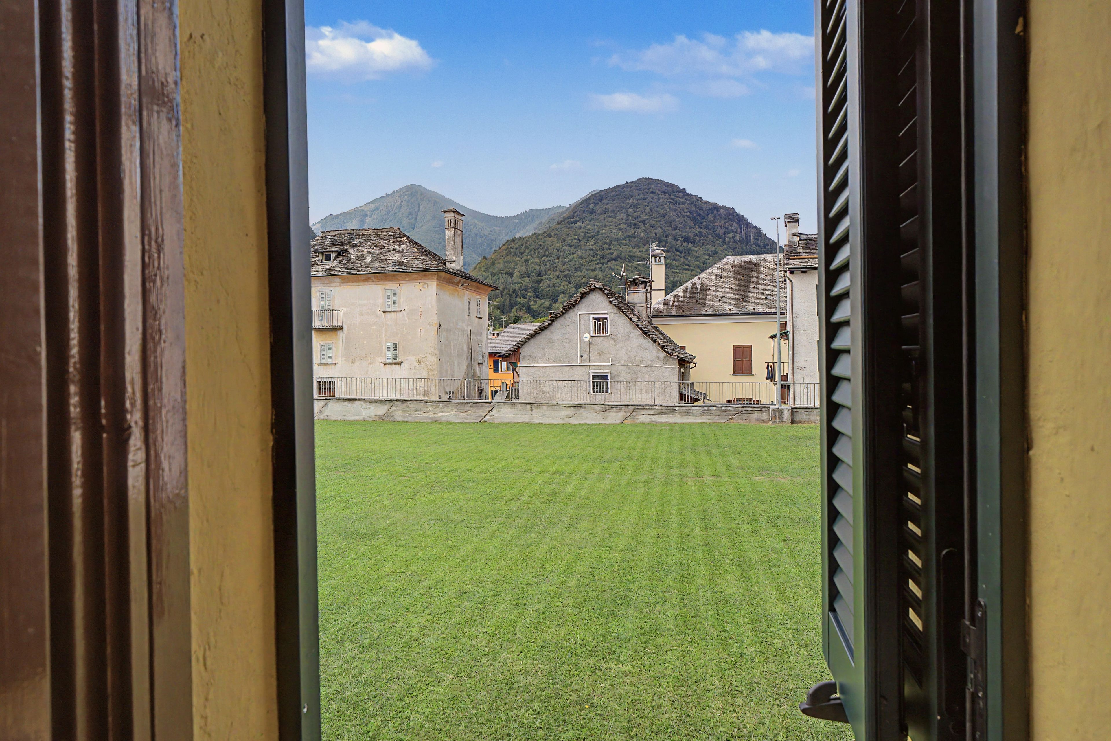 Vista a través de una ventana de un edificio con un patio cubierto de césped, una montaña al fondo y una calle con edificios.
