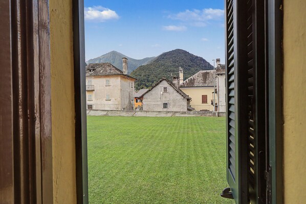 Vista a través de una ventana de un edificio con un patio cubierto de césped, una montaña al fondo y una calle con edificios.