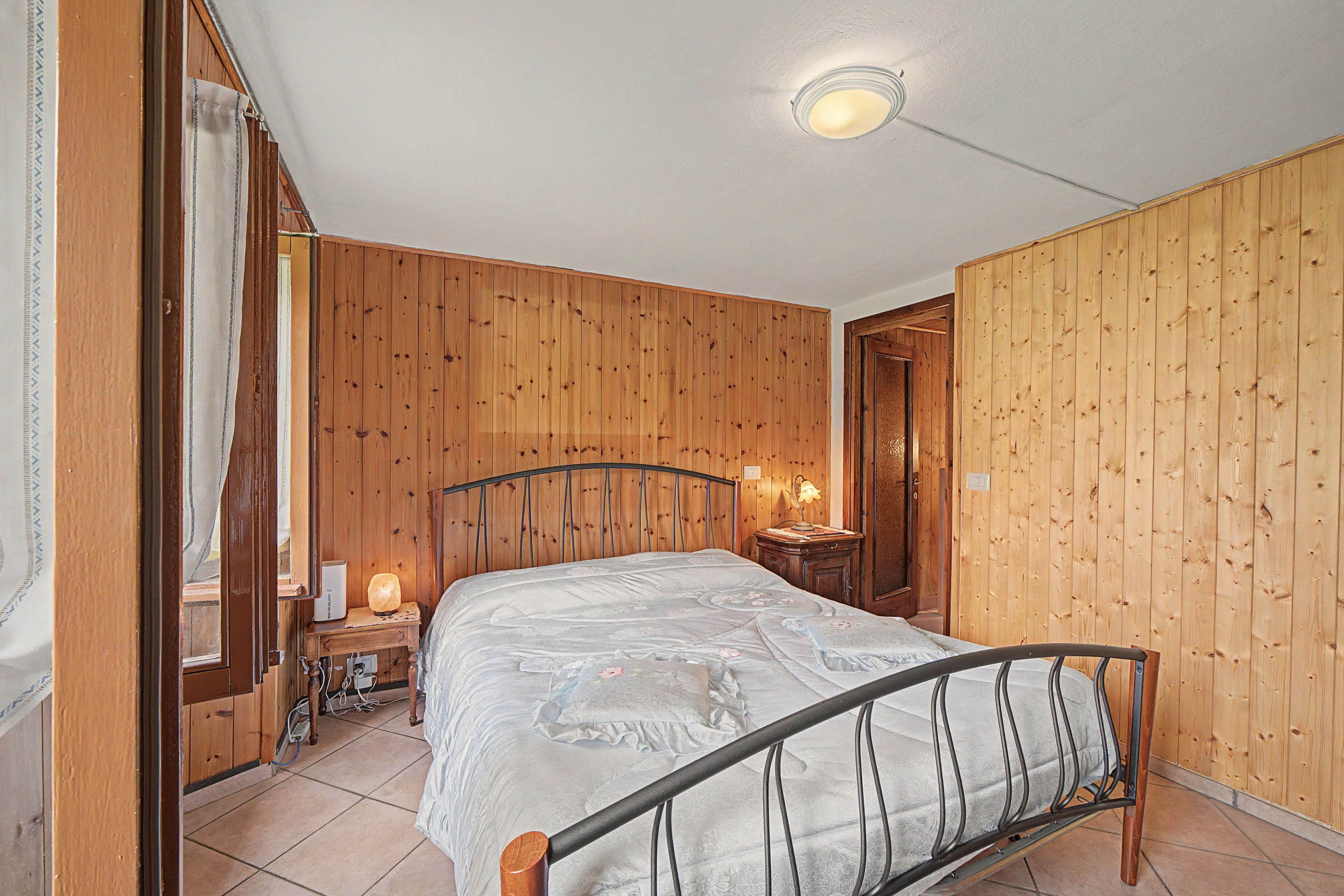 Ein Schlafzimmer mit einer holzverkleideten Wand, einem Bett mit Metallgestell, einem Nachttisch mit Lampe und einer Tür zu einem weiteren Raum.