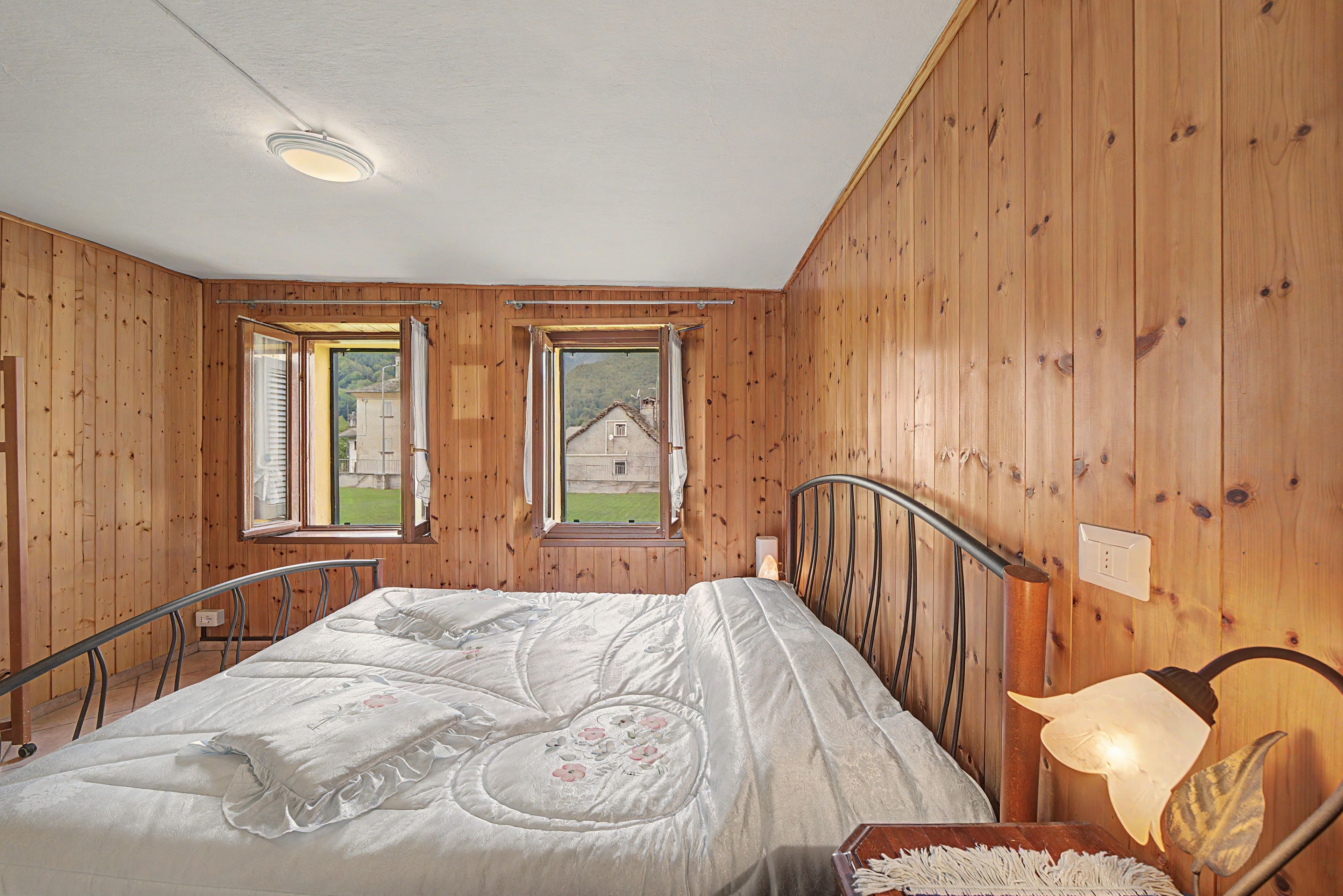 Ein Schlafzimmer mit Holzwänden, einem Bett mit Blumenmuster-Überwurf, zwei Fenstern mit Fensterläden und Blick auf ein Haus.