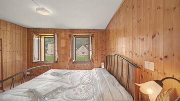 Ein Schlafzimmer mit Holzwänden, einem Bett mit Blumenmuster-Überwurf, zwei Fenstern mit Fensterläden und Blick auf ein Haus.