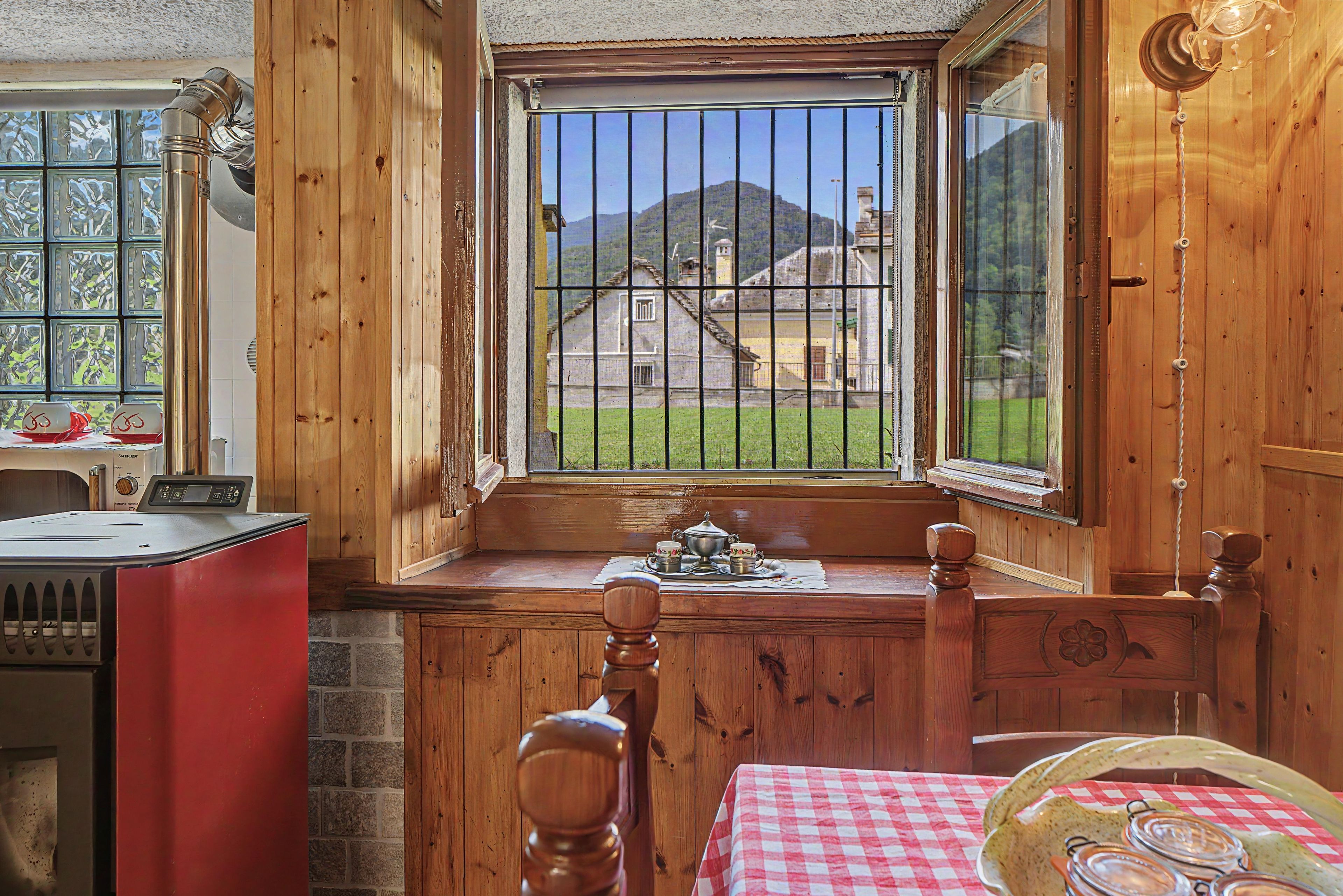 A wooden cabin interior with a kitchen area, a dining table with a checkered tablecloth, and a window overlooking a rural landscape.