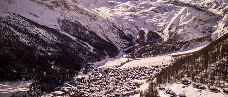 A snow-covered mountain village with buildings clustered along a valley, surrounded by forested slopes and mountains.