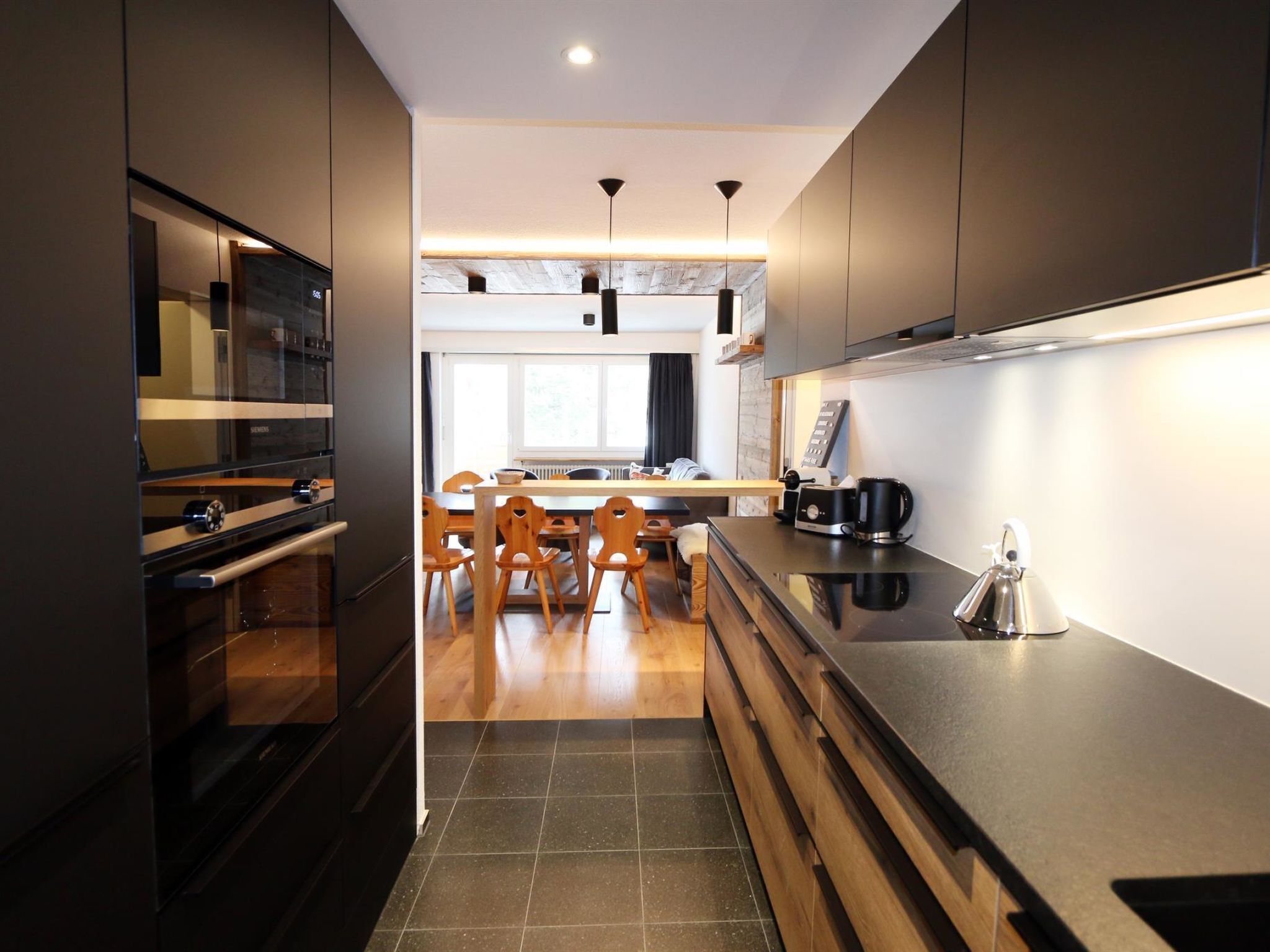 A modern kitchen with dark cabinetry, a central island, and a dining area with wooden chairs.