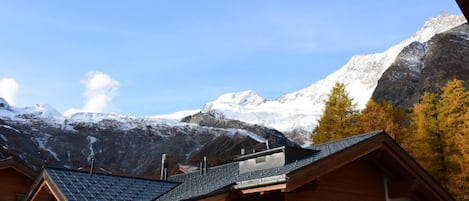 A wooden cabin with a sloped roof, set against a backdrop of snow-capped mountains and clear blue sky.