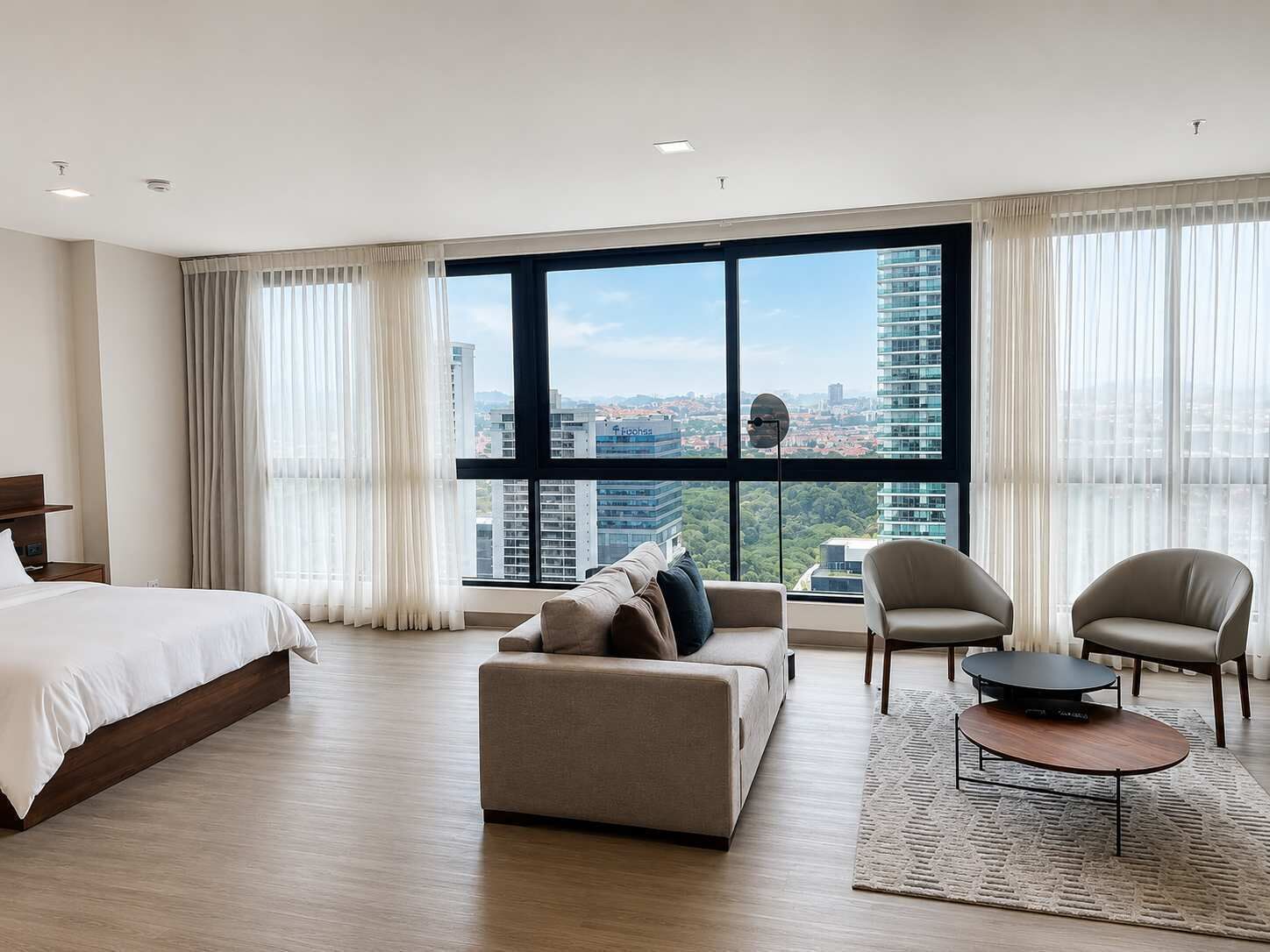 A modern hotel room with a large bed, a sofa, two armchairs, a coffee table, and a view of the cityscape through floor-to-ceiling windows.