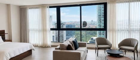 Quarto de hotel moderno com uma cama grande, um sofá, duas poltronas, uma mesa de centro e vista para a paisagem urbana através de janelas do chão ao teto.