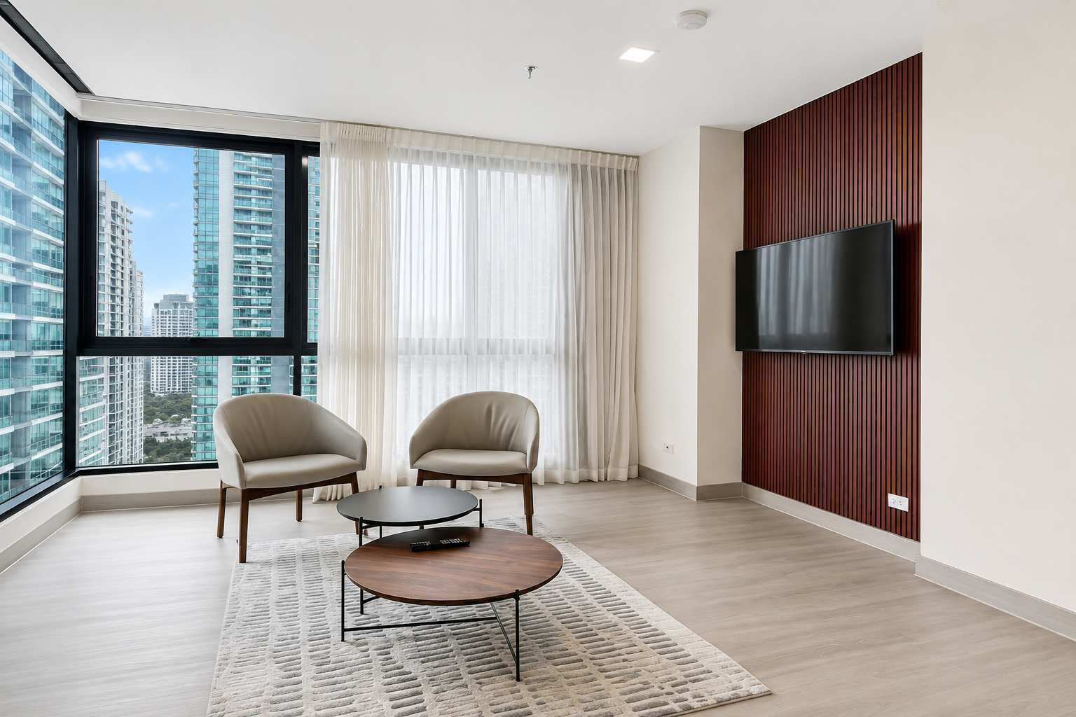 A modern hotel room with a large window, two armchairs, a small round table, and a mounted TV.