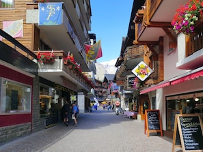Eine enge Straße gesäumt von traditionellen Gebäuden, von denen einige Balkone mit Blumen schmücken, und Schilder, die lokale Geschäfte und Dienstleistungen bezeichnen.