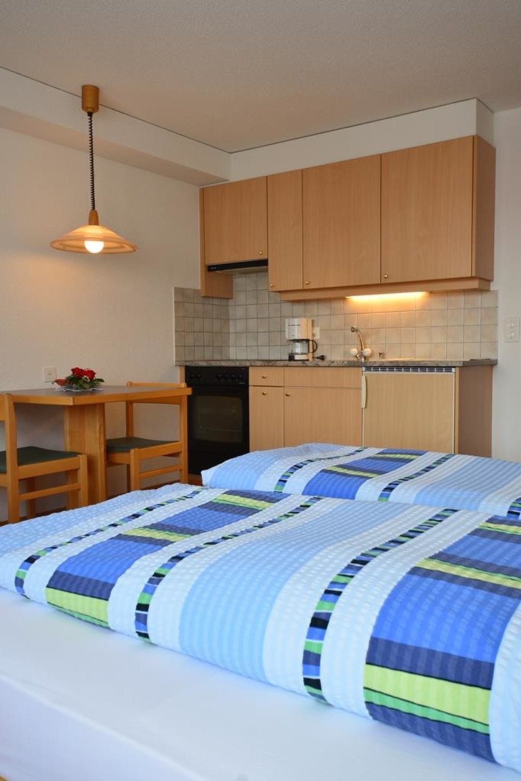 A compact room with a bed, a small table, a chair, a kitchenette, and a hanging light fixture.