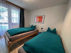 A room with two single beds, a window with curtains, a wall-mounted picture, and a bedside table with a lamp. - Alouette Appartement/Fewo Apartment Lama by Interhome (Saas-Almagell)