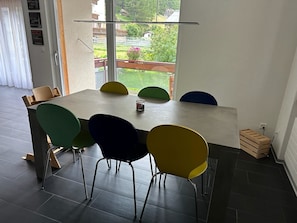 Ein Essbereich mit großem Tisch und bunten Stühlen, ein Fenster mit Blick auf Grünflächen und Gebäude sowie eine kleine Holzkiste auf dem Boden.