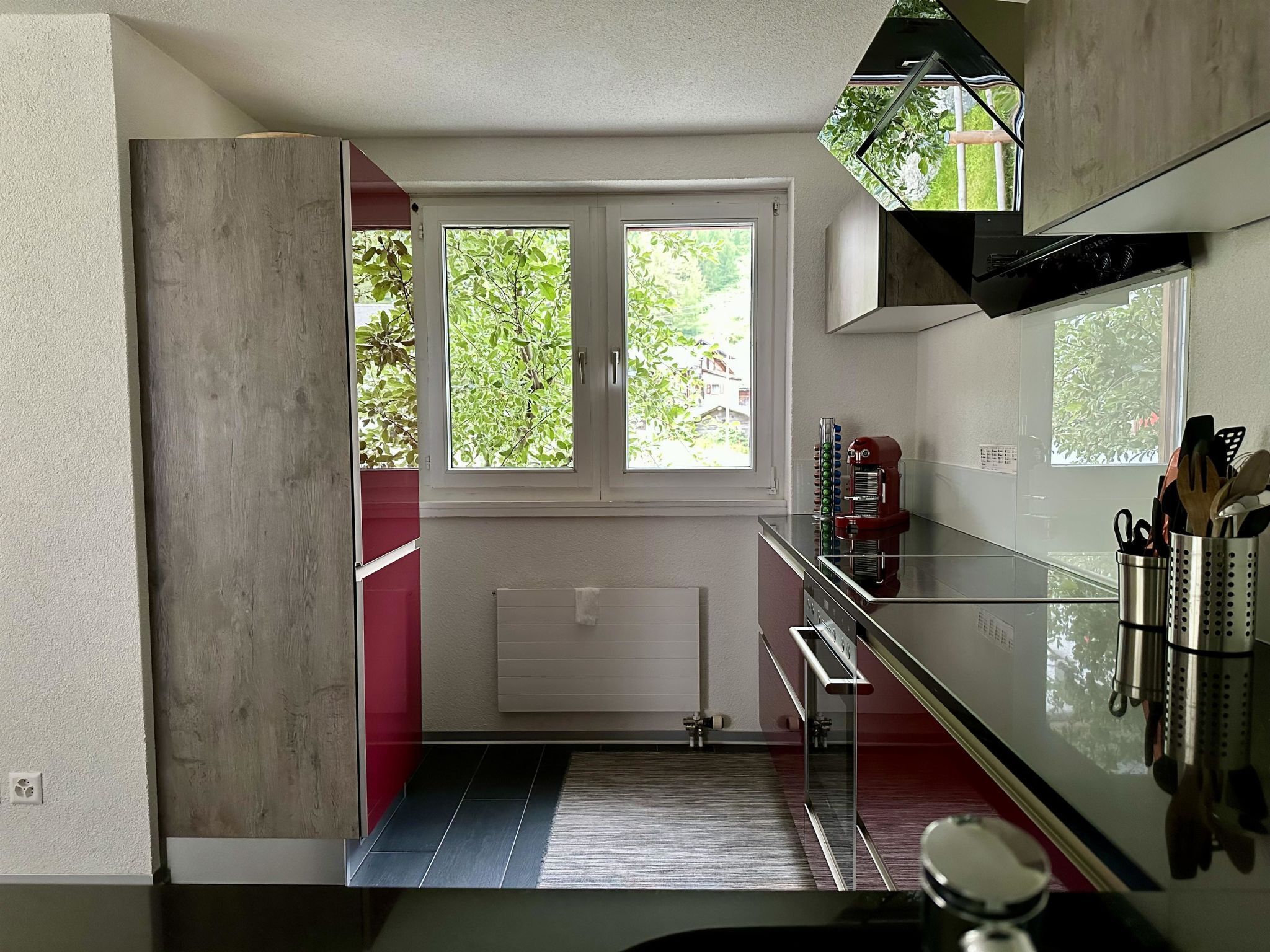 Una cocina moderna con horno empotrado, microondas y encimera de acero inoxidable. Hay dos ventanas que ofrecen luz natural y vistas al exterior.