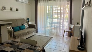 A living room with a sofa, a coffee table, a TV stand, and a view of the outside through a window with curtains. - Apartment near the beach (Menton)