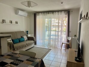 A living room with a sofa, a coffee table, a TV stand, and a view of the outside through a window with curtains. - Apartment near the beach (Menton)
