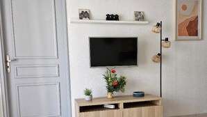 A living room with a television mounted on the wall, a wooden entertainment unit below, a small table with a remote control, and a door to the left. - Apartment near the beach (Menton)
