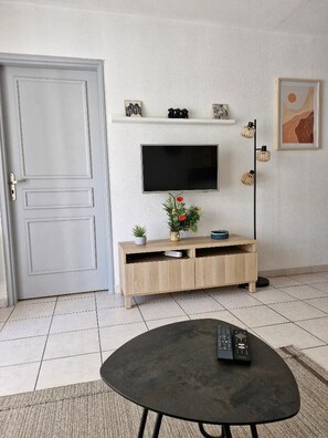 A living room with a television mounted on the wall, a wooden entertainment unit below, a small table with a remote control, and a door to the left. - Apartment near the beach (Menton)