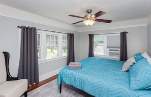 Quarto com cama, cadeira, ventoinha no teto e janelas com cortinas.