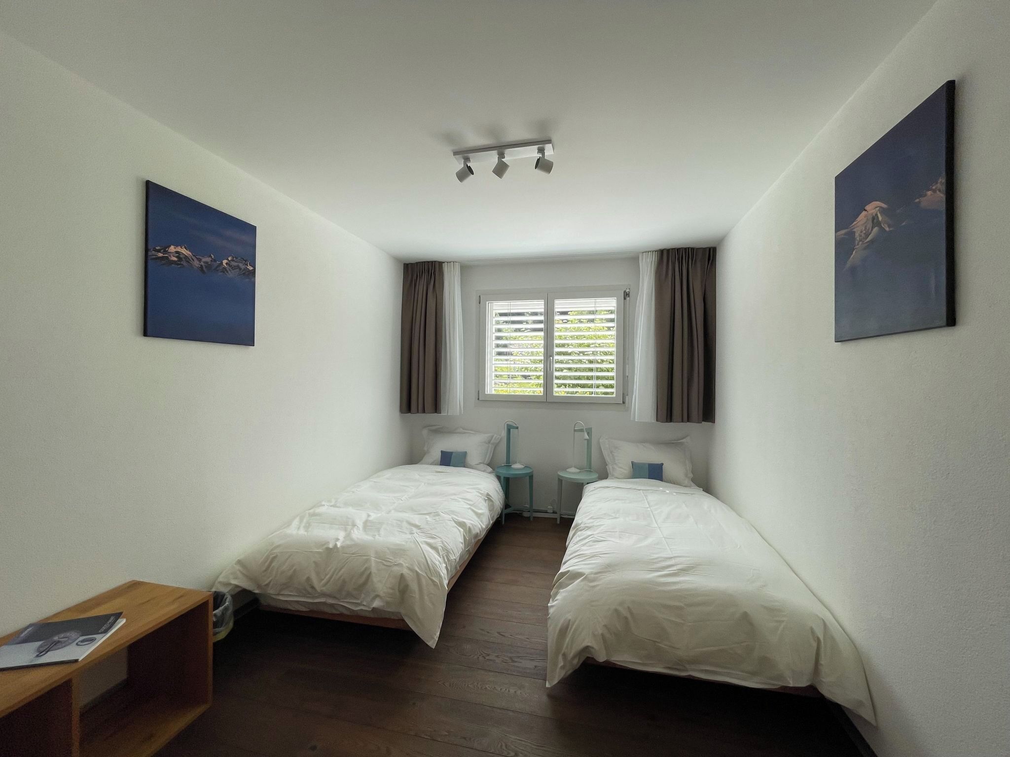 A room with two single beds, a window with blinds, a desk, and two wall-mounted lamps.