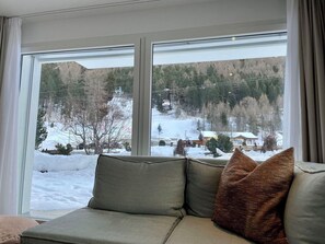 Ein Zimmer mit großem Fenster, einer Couch mit Kissen und Blick auf eine verschneite Landschaft mit Bäumen und einem Gebäude.
