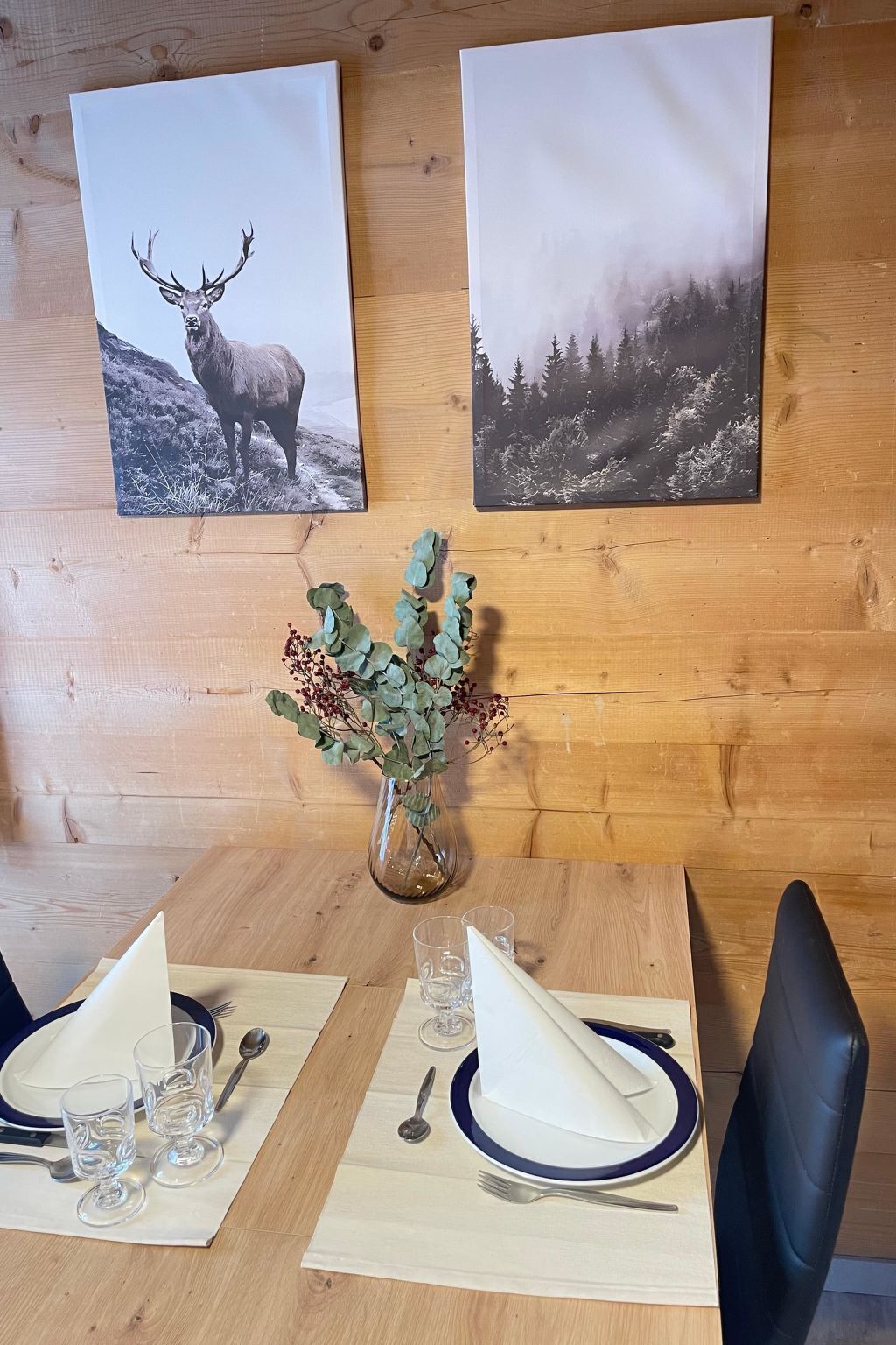 A wooden dining table with two framed wildlife photographs, a vase with green foliage, and a set table with plates, glasses, and cutlery.