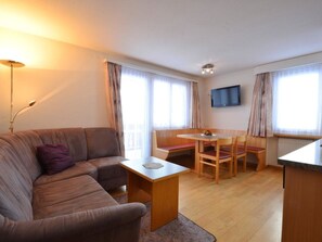 A modern living room with a sofa, a dining table with chairs, a TV mounted on the wall, and large windows with curtains. - Haus Orion Appartement/Fewo 4-Bettwohnung Nr. 6 by Interhome (Saas-Fee)