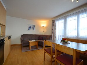 Ein kompakter Wohnbereich mit Küchenzeile, einer Couch, einem Esstisch mit Stühlen und einem großen Fenster mit Vorhängen.